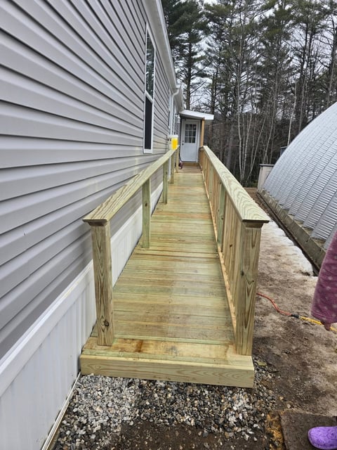 Wooden boardwalk between two buildings with metal siding, bordered by evergreen trees and gravel ground