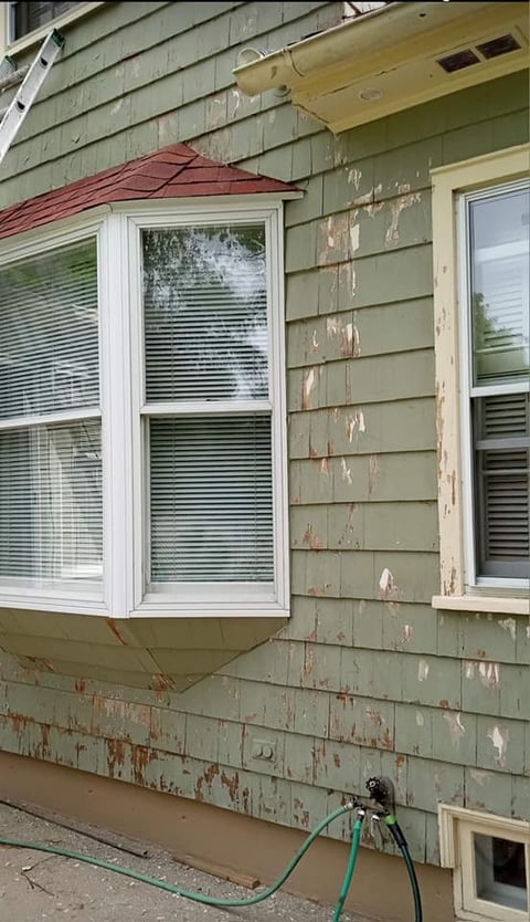 Exterior house wall with peeling green siding, two white windows with blinds, and a red roof awning, with a green hose at the base