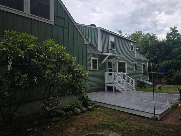 Green two-story house with white railings and deck, surrounded by landscaping and trees under a cloudy sky