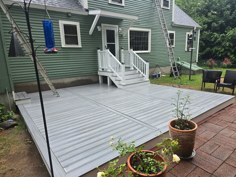 Large white deck attached to green house with potted plants, stairs, and lawn in background