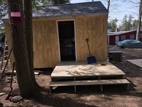 Small wooden shed with dark door and wooden ramp entrance in wooded backyard setting
