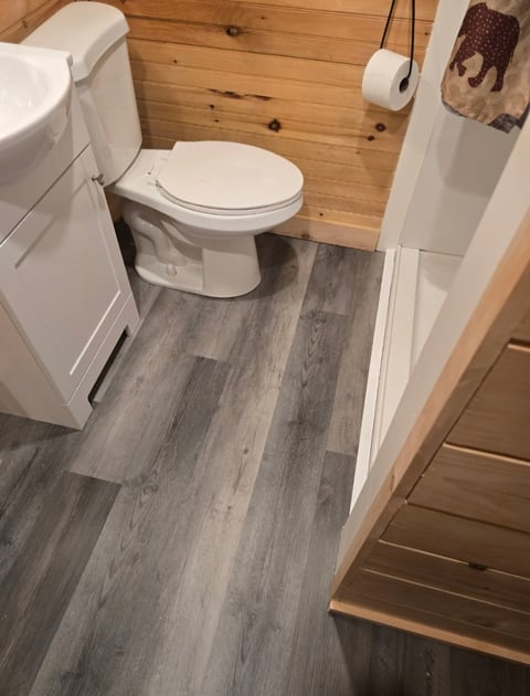 Compact bathroom interior with white toilet, wood-paneled walls, gray wood-grain flooring, and toilet paper holder