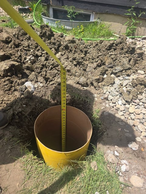 Yellow measuring tape inside a yellow cylindrical pipe set in excavated earth with stones and vegetation visible around the construction site