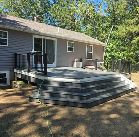 A residential house with a composite deck featuring tiered steps, black railings, and a grill, surrounded by mature trees on a sunny day.