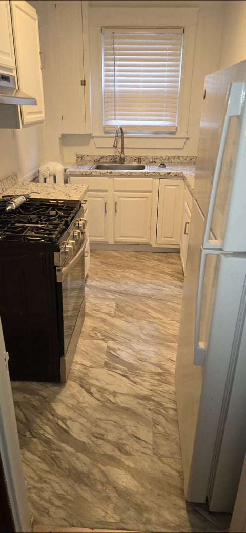 Modern kitchen with white cabinets, granite countertop, black gas stove, stainless steel refrigerator, and wood-look tile flooring with window above sink