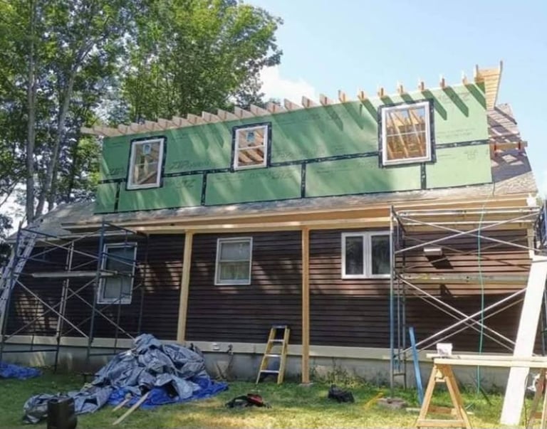 House under renovation with exposed framing, green roof tarps, scaffolding, and construction materials in yard