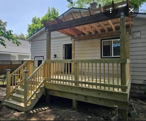 Single-story house with wooden deck, pergola, and railings on a sunny day with green trees visible