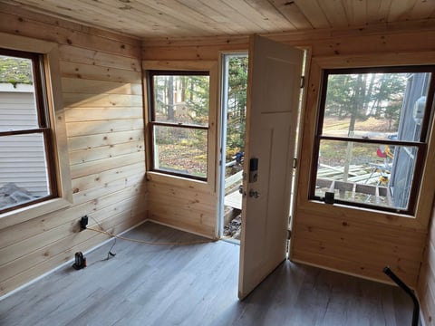 Interior of a wooden cabin with open door, windows showing forest views, and gray laminate flooring