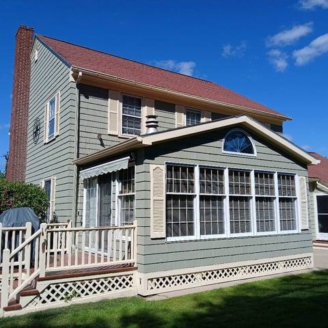 Two-story white and cream colonial house with red roof, brick chimney, front porch with railings, and multiple windows under blue sky