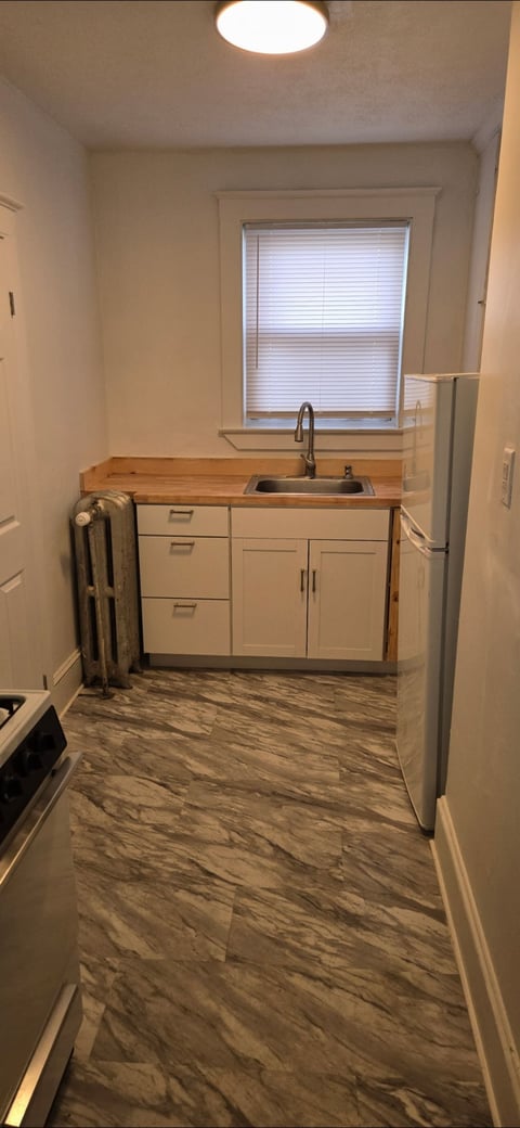 Small kitchen with white cabinets, wooden countertop, sink, window with blinds, and wood-grain vinyl flooring