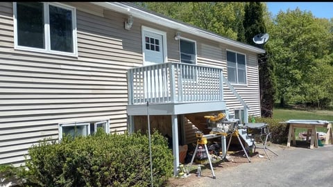Gray vinyl-sided single-story house with white railings on an elevated deck, landscaped with bushes and trees, with vehicles and equipment underneath