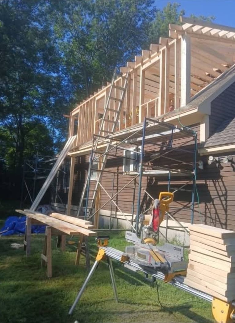 Home addition under construction with exposed wooden framing, metal scaffolding, and tools on the grass