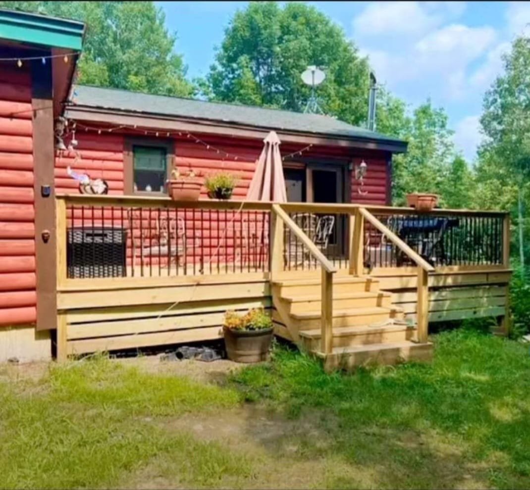 Red log cabin with wooden deck and railings surrounded by green trees and lawn