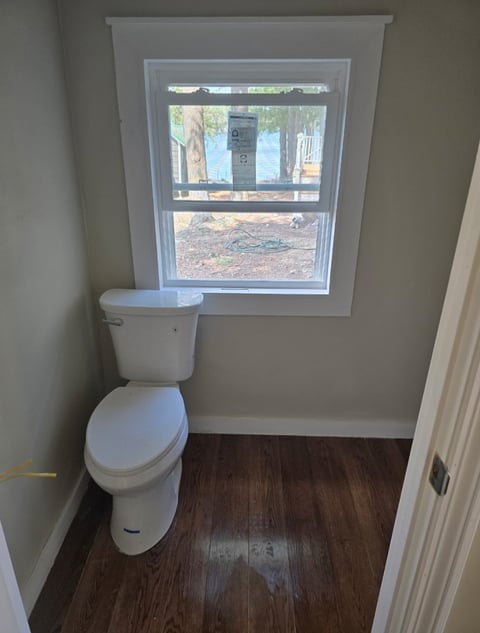 A white toilet in a small bathroom with a window on the wall and dark wood flooring