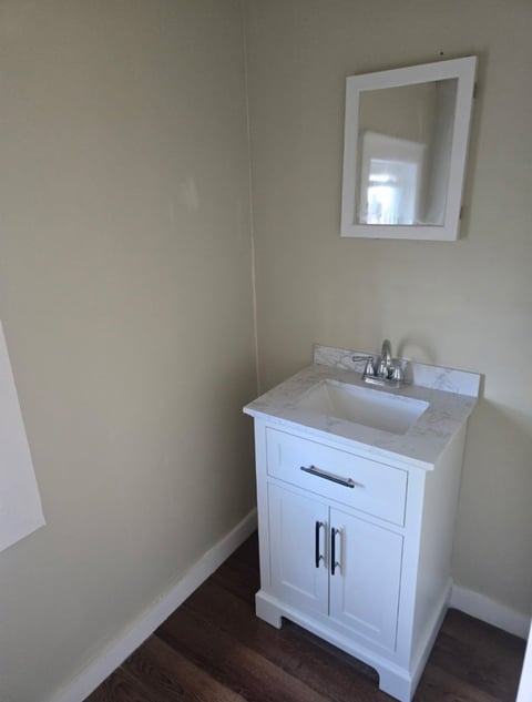 White bathroom vanity with sink and faucet, white framed mirror on wall above, in a corner with beige walls and dark wood flooring