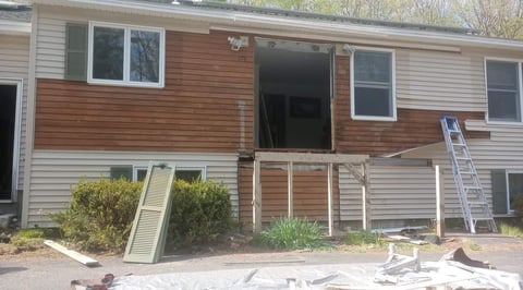 Damaged multi-level residential home with boarded windows, wooden deck, and construction materials in foreground