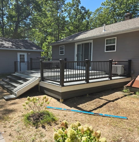 Single-story gray house with black deck railing, wooden steps, and landscaped yard with blue hose