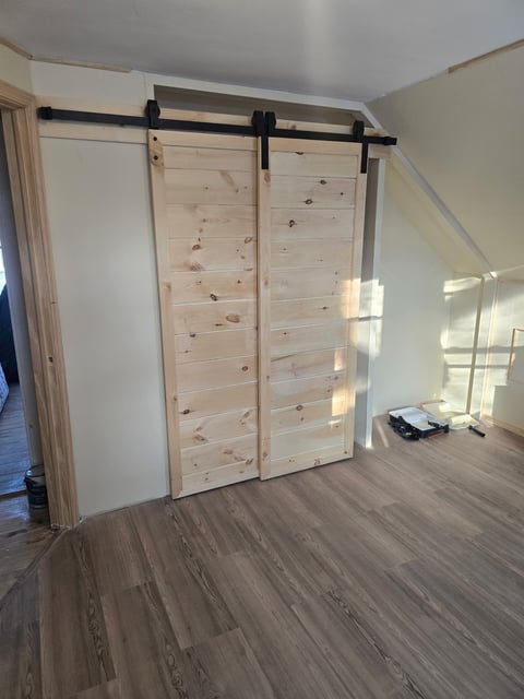 Light wood barn doors with black hardware mounted on ceiling track in bright, minimalist room with wood flooring