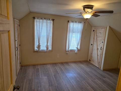Empty room with wood plank flooring, beige walls, two windows with blinds, ceiling fan light fixture, and white interior doors