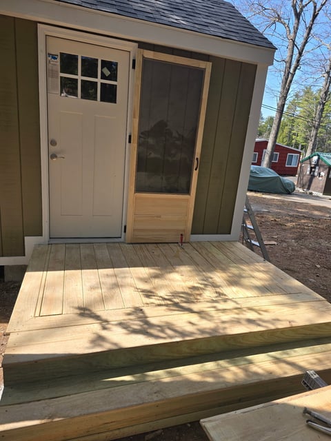 Small green shed with wooden deck, beige door with window, and screen door entrance