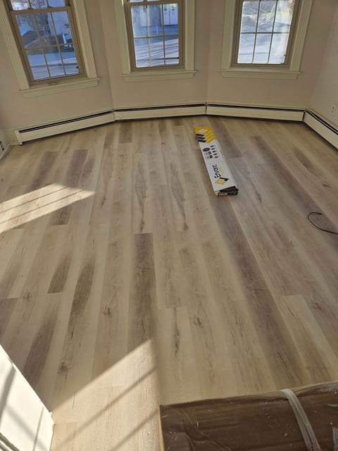 Empty room with light wood laminate flooring, black baseboard trim, three windows, and construction materials on floor
