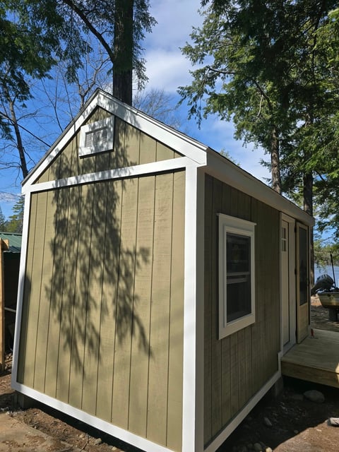 Small tan and white shed with pitched roof surrounded by trees on a sunny day